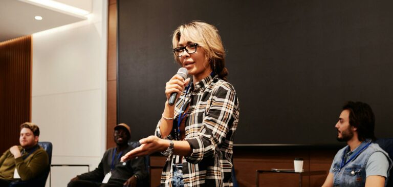 A confident woman speaks into a microphone during a panel discussion indoors.