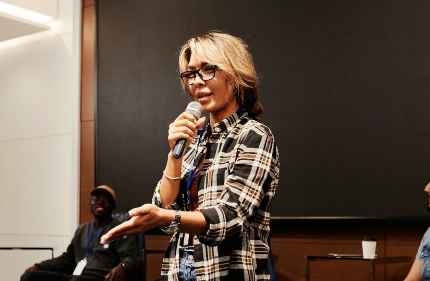 A confident woman speaks into a microphone during a panel discussion indoors.
